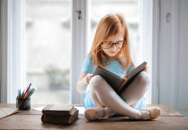 petite fille rousse avec des lunettes qui lit sur une table en bois